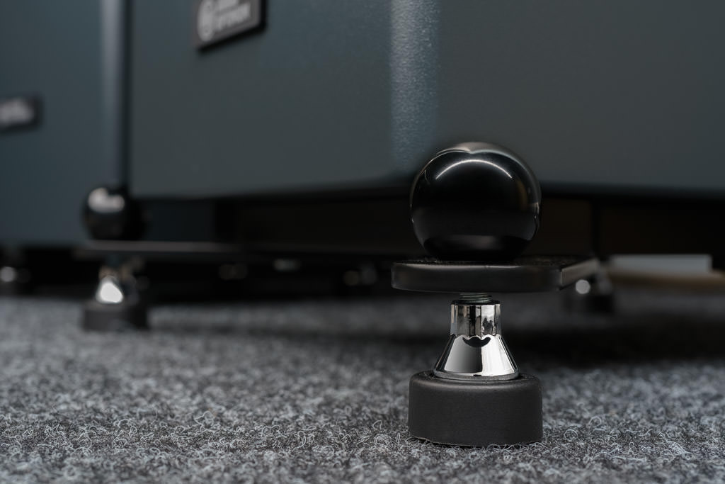 Die Lautsprecher sind auf Auslegern gelagert. Dies erhöht die Standfestigkeit, befördert aber auch eine leichtere Anmutung: Das Gehäuse scheint zu schweben. Zur Minimierung von Vibrationen sitzt zwischen Korpus und Auslegern eine absorbierende Unterfütterung. Die Füße sind höhenverstellbar, die zierende oberseitig aufgeschraubte Kugel dient als fixierende Konterung. Bei den Füßen handelt es sich um die von Audio Optimum stets eingesetzten Soundcare Superspikes. Sie bewirken eine Entkopplung und passen sich unebenen Untergründen an.
