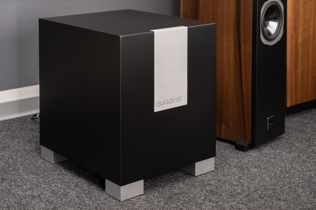 Der wahlweise in hochglänzendem Schwarz oder Weiß gehaltene Subwoofer Qube CS10 erweist sich mit seinen gerundeten Kanten, der gebürsteten Aluminium-Applikation und den stimmig gestalteten Füßen als attraktiver Tiefton-Lieferant. Die Füße sorgen für einen definierten Bodenabstand, damit der Schall des gen Untergrund gerichteten Woofers in alle Richtungen gleichmäßig abgestrahlt werden kann. Auch der Bassreflex-Kanal verläuft über die Front in Richtung Boden. Der Kanal ist strömungsoptimiert, so entstehen weniger Ventilationsgeräusche. Im Test ist von der bewegten Luft dementsprechend nichts zu hören.