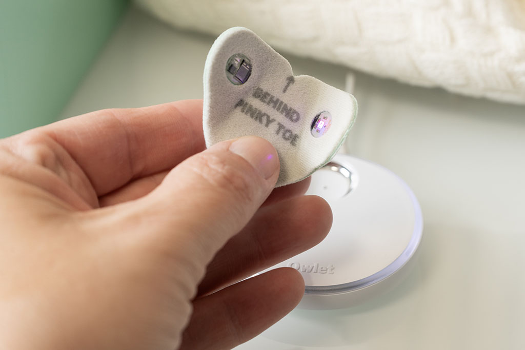 Hier sehen wir die Innenseite des einsetzbaren Sensors: rechts sitzt der Sender, der Licht durch den Fuß strahlt, links der Detektor. Mithilfe der Durchleuchtung werden Herzfrequenz, Sauerstoffsättigung und Baby-Bewegung ermittelt. Diese Daten werden über den ebenfalls integrierten Bluetooth-Sender an die Basisstation gesendet. Die Technik ist so in den Kunststoff der Sensor-Einheit eingelassen, dass sie ohne Kontakt mit der Haut des Kindes ist.