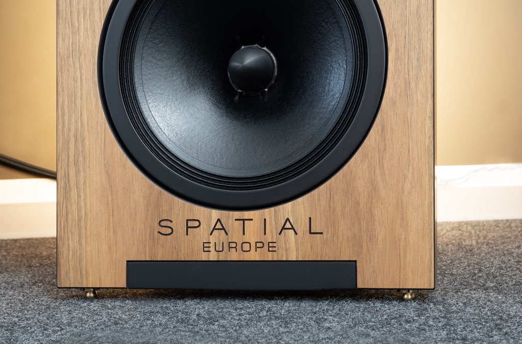 Der eingraviert Firmen-Schriftzug setzt den schwarzen Kontrast der Schallwand-Kanten, der Chassis-Rahmungen und des Metallfußes fort. Dieser Fuß ist durch die Schallwand geführt, wo er frontseitig bündig mit ihr abschließt. Schallwand und Fuß sind zudem verschraubt, der daraus resultierende definierte Anpressdruck reduziert Vibrationen.