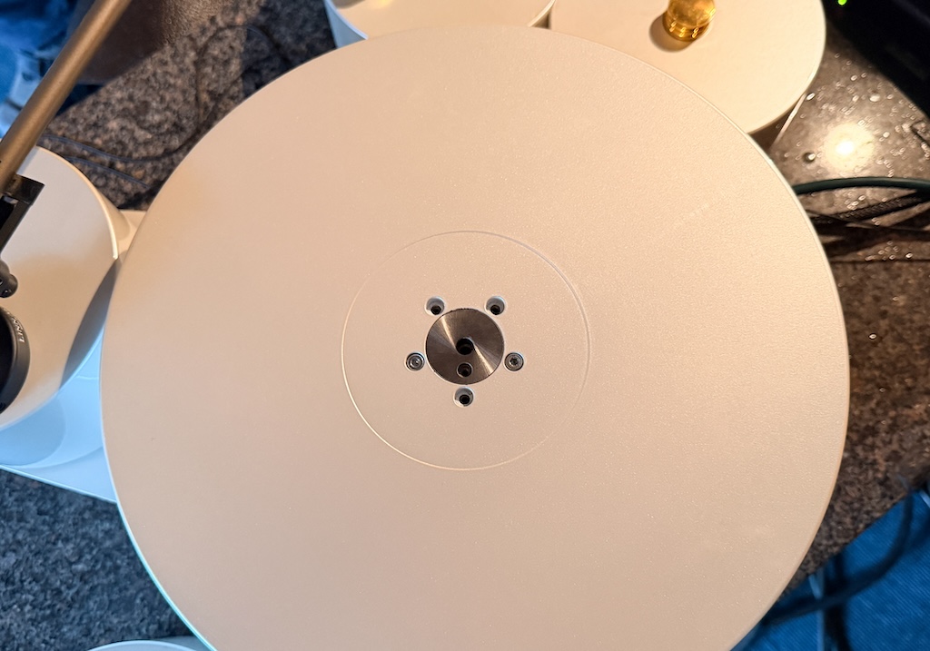 Der Teller ist normalerweise mit der mitgelieferten Spezial-Matte aus Rindsleder belegt. Ohne Auflage sieht man die Verschraubung des 21-Kilo-Tellers mit der Subteller-artigen Hülse des Lagers. Mit ihr ist der Teller hier nur über zwei Schrauben verbunden. Das Lager ist derart sensibel, dass jede weitere Schraube klangliche Veränderungen nach sich zieht. Der obere separate Teil der Spindel ist abnehmbar. Durch die Teilung bleibt auch nach dem Einsetzen die energetische Verbindung zum Lager unterbrochen.